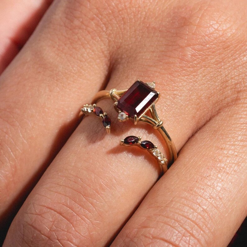 May include: A gold ring featuring a large, rectangular, deep red gemstone as the focal point. The ring has a split shank design, with smaller red and white gemstones accenting the band. The ring is set on a finger.