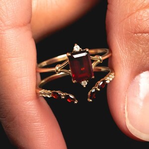 May include: A gold ring with a large, rectangular, deep red gemstone as the centerpiece. The ring has a delicate design with smaller red gemstones and clear accent stones. The ring is held against a dark background.