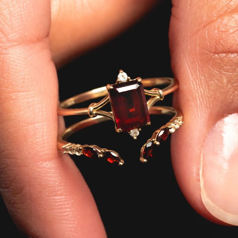 May include: A gold ring with a large, rectangular, deep red gemstone as the centerpiece. The ring has a delicate design with smaller red gemstones and clear accent stones. The ring is held against a dark background.