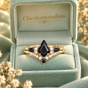 May include: A gold ring set featuring a central kite-shaped black gemstone, accented by smaller marquise-cut black stones and round white stones in a chevron band. The ring is displayed inside a sage green velvet box with Chiccharmstudious text.
