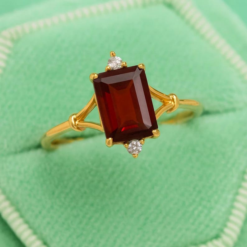 May include: A gold ring featuring a rectangular, deep red gemstone with two small, round, clear accent stones. The ring has a delicate, openwork design and is set against a soft green background.