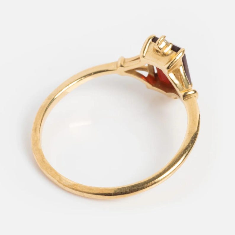 May include: A gold ring with a rectangular, dark red gemstone. The ring has a thin band and a detailed setting around the stone. The setting has a vintage style with decorative elements.