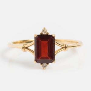 May include: A gold ring featuring a rectangular, deep red gemstone. Two smaller, round, clear gemstones are set above and below the main stone. The ring has a delicate, vintage design.
