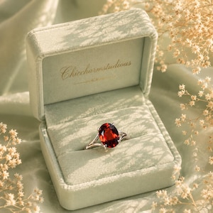 May include: A silver ring featuring a large, oval-cut deep red gemstone set in a prong setting. The ring is displayed inside a light sage green velvet jewelry box with the text Chiccharmstudious printed in gold on the inner lid lining.