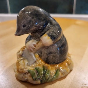 May include: A ceramic mole figurine, shown seated and holding a shovel. The mole is predominantly grey and black, with tan and cream details. It rests on a mound of tan and green-coloured earth, typical of garden ornaments.