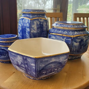 May include: A set of blue and white ceramic tableware with gold edging. The collection includes a square bowl, two lidded jars, and a larger jar. Each piece features a blue illustration of a bridge or building.