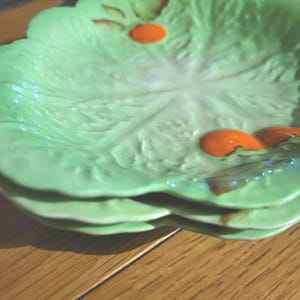 May include: Three light green ceramic plates, each shaped like a lettuce leaf. The plates have a textured surface with orange accents and brown details. The plates are stacked on a wooden surface.