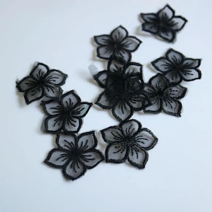 Puede incluir: Un conjunto de ocho apliques individuales con forma de flor negra. Cada pieza presenta un centro gris translúcido y transparente con bordes bordados en negro detallados y venas de pétalos radiantes, creando un delicado motivo floral textil.
