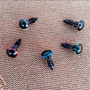 May include: A collection of black plastic safety eyes with colored washers in red, blue, and green. The eyes have a glossy, reflective surface and are designed for crafts and toy making. The background is a textured, brown surface.