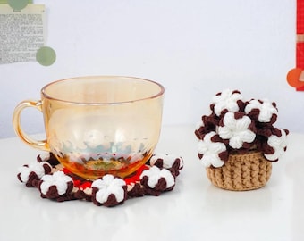 Könnte beinhalten: Eine klare Glastasse steht auf einem rot-weiß gehäkelten Untersetzer mit braunen und weißen Blumenakzenten. Ein kleiner gehäkelter Korb enthält einen Strauß weißer und brauner gehäkelter Blumen. Die Artikel befinden sich auf einer weißen Oberfläche.