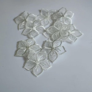 May include: A collection of six white floral lace appliqués, each featuring five petals with delicate embroidered edges and fine stitching details radiating from the centre, arranged in a scattered pattern on a plain white surface.