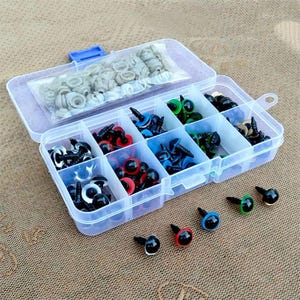 May include: A clear plastic organizer filled with various sizes and colors of craft safety eyes. The eyes are black, red, blue, and green, with black stems. A small bag of gray washers is in the top compartment. The organizer is on a brown surface.