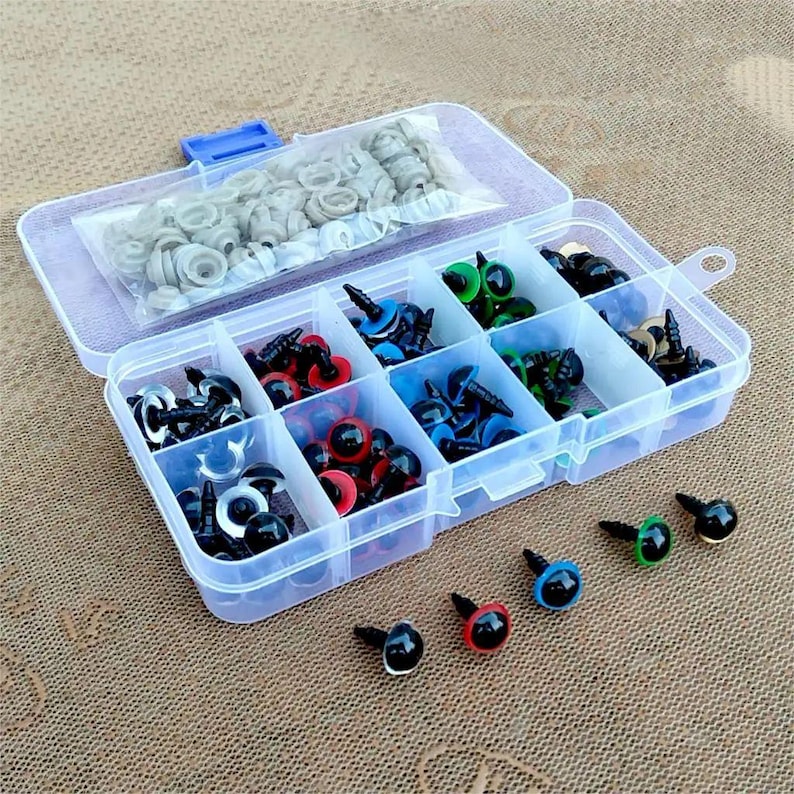 May include: A clear plastic organizer filled with various sizes and colors of craft safety eyes. The eyes are black, red, blue, and green, with black stems. A small bag of gray washers is in the top compartment. The organizer is on a brown surface.
