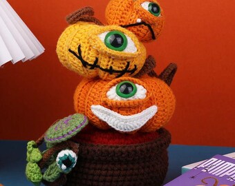 May include: A stack of three crocheted pumpkins with large green eyes and smiling faces. The pumpkins are orange and yellow, with brown stems and black stitched details. The base is a brown crocheted pot with a green and purple creature.