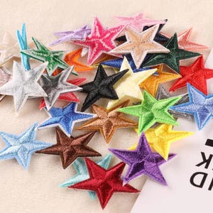 May include: A selection of embroidered star patches in a variety of colours, such as red, blue, green, and gold. The patches are scattered, each star featuring a detailed, stitched design. These patches are suitable for crafting and adding decorative flair to items.