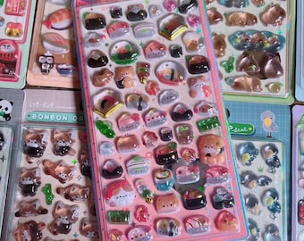 May include: A collection of colorful sticker sheets featuring various designs. One sheet showcases sushi-themed stickers, while others display animal and food-related graphics. The stickers are in a variety of shapes and sizes, with a glossy finish.