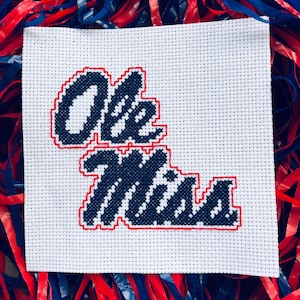 May include: A white cross-stitch fabric square with the words "Ole Miss" in navy blue, outlined in red. The fabric is surrounded by red, white, and blue decorative streamers.