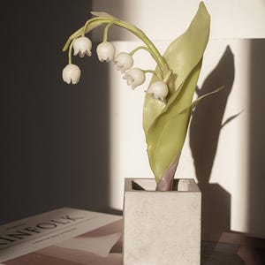 Puede incluir: Un arreglo floral decorativo con flores blancas en forma de campana y una gran hoja verde. Las flores están en una maceta pequeña y cuadrada, similar al hormigón. El arreglo está sobre una mesa junto a un libro.