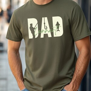 May include: Olive green t-shirt with a graphic design featuring the word "DAD" in large, off-white letters. The word "Tennant" is written in a green cursive font. The design includes a silhouette of a fisherman, a fish hook, and a fish.