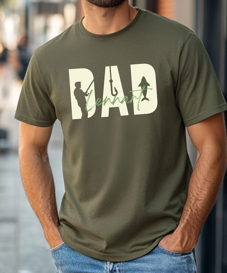 May include: Olive green t-shirt with a graphic design featuring the word "DAD" in large, off-white letters. The word "Tennant" is written in a green cursive font. The design includes a silhouette of a fisherman, a fish hook, and a fish.