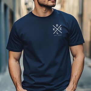 May include: Dark navy blue crew neck t-shirt with a white graphic on the chest. The graphic features the letters "RKE" in a cross design, with "EST. 2023" below. The shirt is made of a soft material.