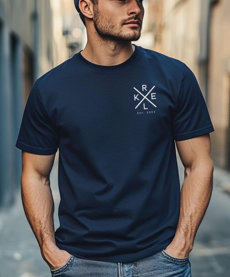 May include: Dark navy blue crew neck t-shirt with a white graphic on the chest. The graphic features the letters "RKE" in a cross design, with "EST. 2023" below. The shirt is made of a soft material.