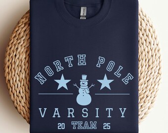 May include: A navy blue sweatshirt featuring the text "NORTH POLE VARSITY TEAM 25" in light blue, with star and snowman graphics. The garment is displayed on a woven surface, suitable for winter wear.