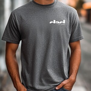 May include: A dark gray t-shirt with the white text "dad of one" printed on the upper left chest. The shirt is short-sleeved and made of a soft material. The person is wearing blue jeans.