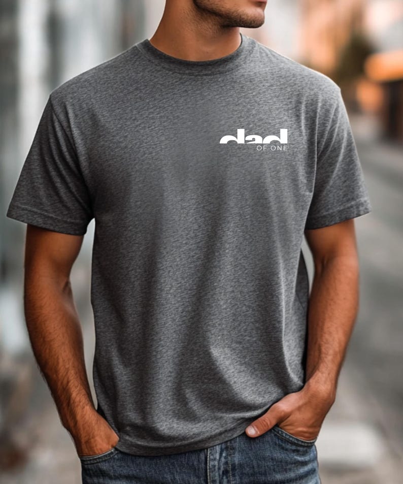 May include: A dark gray t-shirt with the white text "dad of one" printed on the upper left chest. The shirt is short-sleeved and made of a soft material. The person is wearing blue jeans.