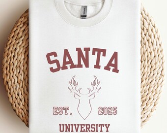 May include: A white sweatshirt featuring the word "SANTA" in maroon block letters above a deer head graphic. Below the graphic are the words "EST. 2025 UNIVERSITY" in maroon. The sweatshirt is folded on a woven, circular surface.
