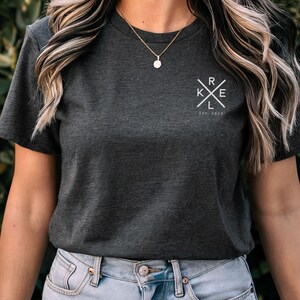 May include: Charcoal grey t-shirt featuring a white graphic on the upper left chest. The graphic is a cross with the letters "RKE" inside and the text "Est. 2022" below. The model is wearing a gold necklace and blue jeans.