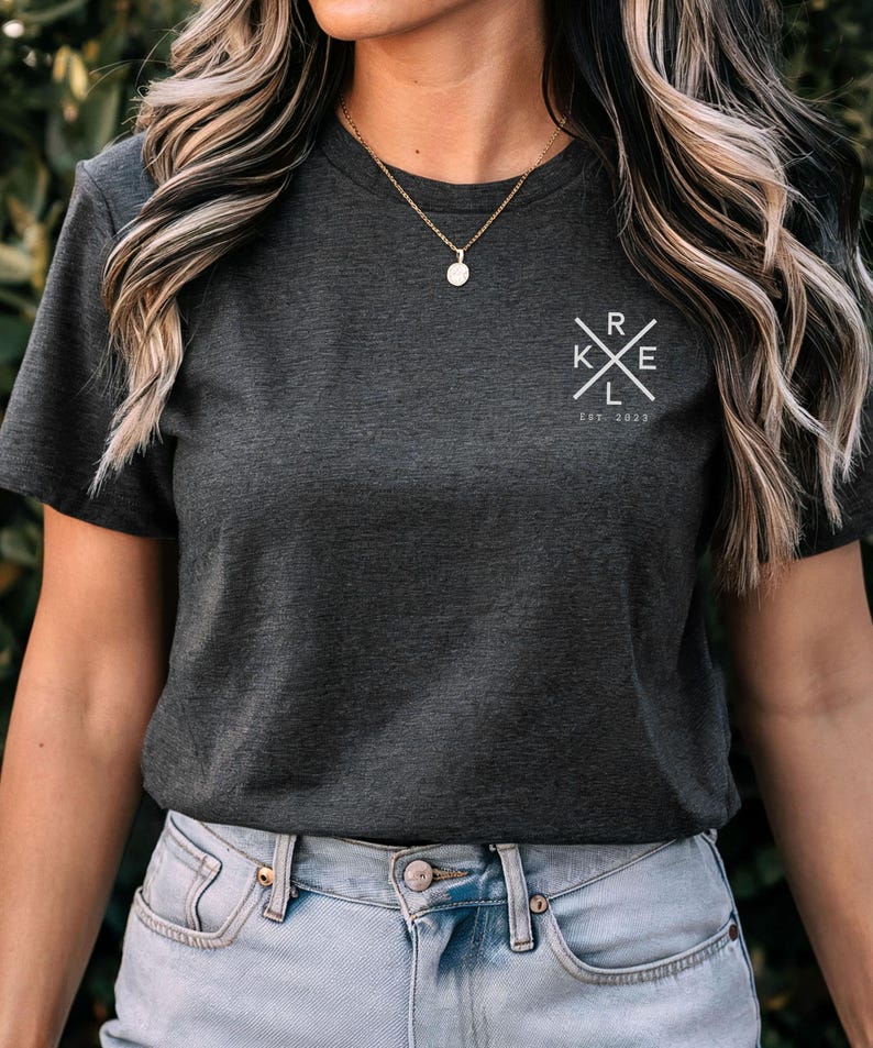 May include: Charcoal grey t-shirt featuring a white graphic on the upper left chest. The graphic is a cross with the letters "RKE" inside and the text "Est. 2022" below. The model is wearing a gold necklace and blue jeans.