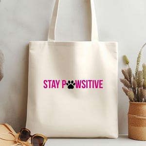 Pu&ograve; includere: Borsa tote color crema con la scritta rosa "STAY PAWSITIVE" e un'impronta di zampa nera. La borsa ha manici lunghi. Sullo sfondo, occhiali da sole e un cestino di vimini con fiori secchi.