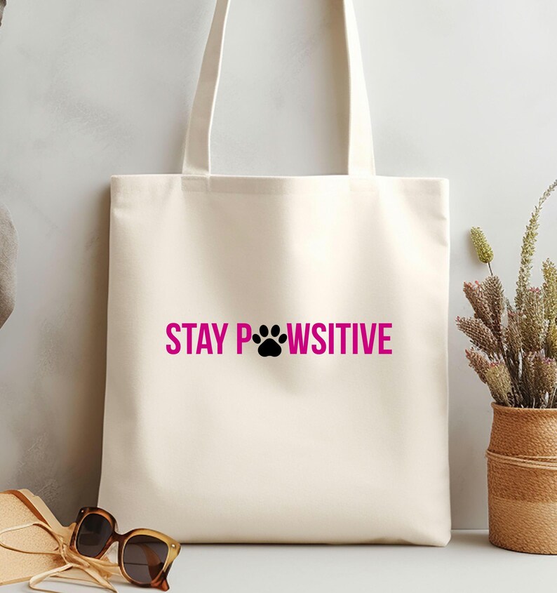 Pu&ograve; includere: Borsa tote color crema con la scritta rosa "STAY PAWSITIVE" e un'impronta di zampa nera. La borsa ha manici lunghi. Sullo sfondo, occhiali da sole e un cestino di vimini con fiori secchi.