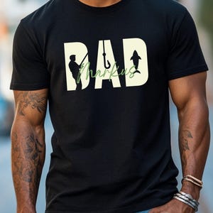 May include: Black t-shirt with a fishing-themed design. The word "DAD" is in a large, off-white font, with the name "Markus" in a green, cursive font. A silhouette of a fisherman, a fish hook, and a shark are incorporated into the design.