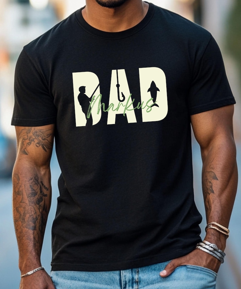 May include: Black t-shirt with a fishing-themed design. The word "DAD" is in a large, off-white font, with the name "Markus" in a green, cursive font. A silhouette of a fisherman, a fish hook, and a shark are incorporated into the design.
