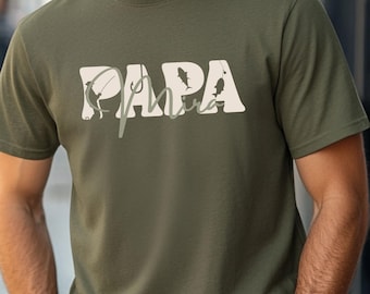May include: An olive green t-shirt featuring the word "PAPA" in a blocky, off-white font. The letter "A" is stylized with a fishing hook and fish. The word "Papa" is overlaid with the word "Mira" in a cursive font.