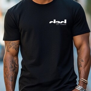May include: Black crew neck t-shirt with the white text "dad of three" printed on the upper left chest. The shirt is made of a soft material and has short sleeves.