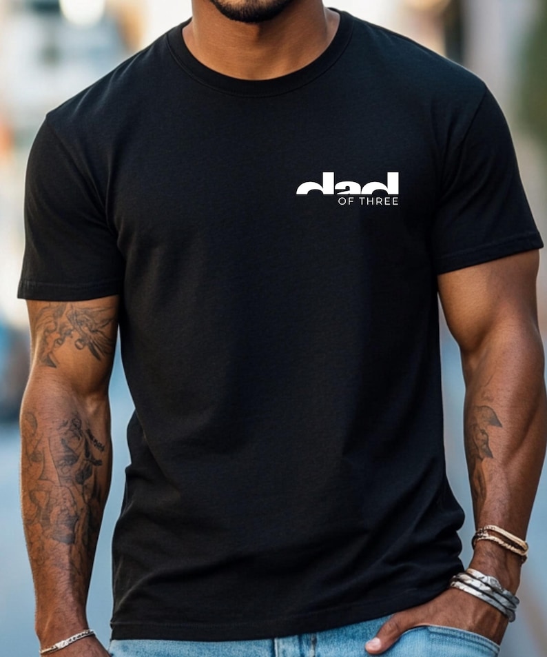 May include: Black crew neck t-shirt with the white text "dad of three" printed on the upper left chest. The shirt is made of a soft material and has short sleeves.