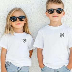 May include: Two children in white t-shirts and sunglasses. Each shirt features a black graphic with the letters R, K, E, and L, and the text "Est. 2023". They are standing against a white wall.
