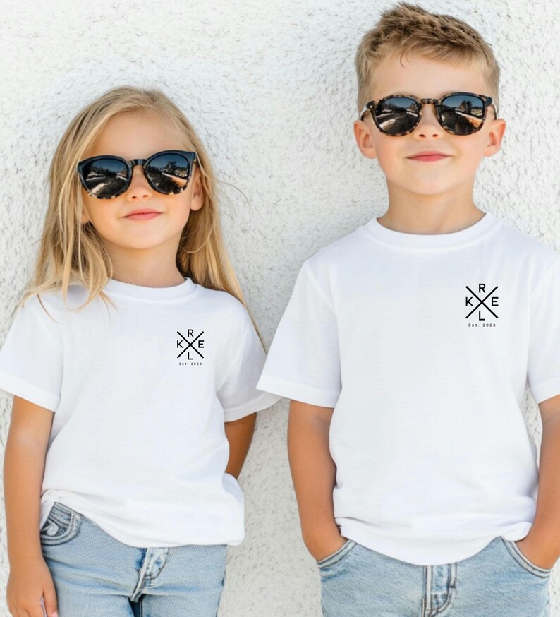 May include: Two children in white t-shirts and sunglasses. Each shirt features a black graphic with the letters R, K, E, and L, and the text "Est. 2023". They are standing against a white wall.