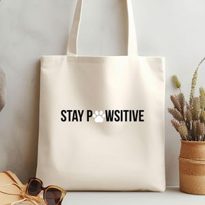 Pu&ograve; includere: Borsa tote color crema con la scritta "STAY PAWSITIVE" in nero, con un'impronta di zampa al posto della "O". La borsa ha due manici ed &egrave; realizzata in tessuto colorato chiaro. Occhiali da sole e un cappello di paglia in primo piano.