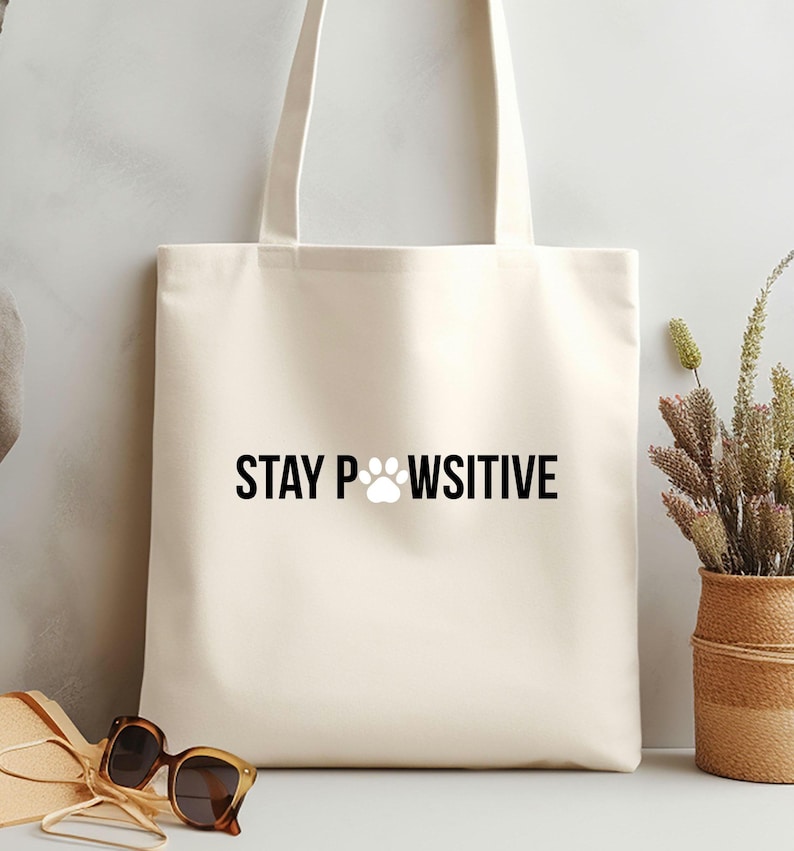 Pu&ograve; includere: Borsa tote color crema con la scritta "STAY PAWSITIVE" in nero, con un'impronta di zampa al posto della "O". La borsa ha due manici ed &egrave; realizzata in tessuto colorato chiaro. Occhiali da sole e un cappello di paglia in primo piano.