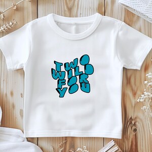 Könnte beinhalten: Weißes T-Shirt mit dem Text "TWO WILD FOR YOU" in blauen Buchstaben mit schwarzer Umrandung. Das Shirt wird auf einer Holzoberfläche präsentiert.