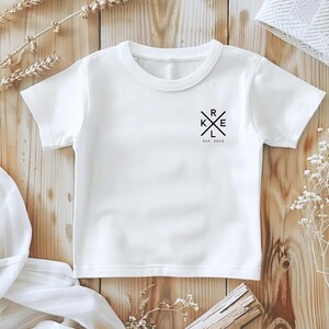 May include: A white t-shirt with a crew neck and short sleeves. The shirt has a black graphic design on the upper left chest with the letters K, R, E, and L in a cross shape, with the text "Est. 2023" underneath.