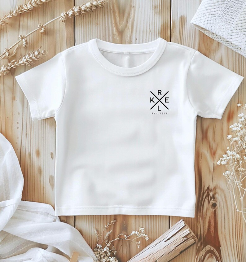 May include: A white t-shirt with a crew neck and short sleeves. The shirt has a black graphic design on the upper left chest with the letters K, R, E, and L in a cross shape, with the text "Est. 2023" underneath.