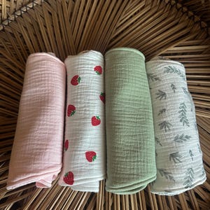 May include: Four rolled muslin cloths in a woven basket. One is light pink, one is white with red strawberries, one is light green, and one is white with gray leaf patterns. The cloths are soft and textured.