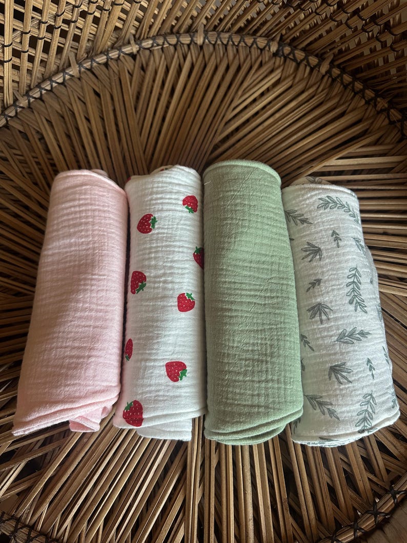 May include: Four rolled muslin cloths in a woven basket. One is light pink, one is white with red strawberries, one is light green, and one is white with gray leaf patterns. The cloths are soft and textured.