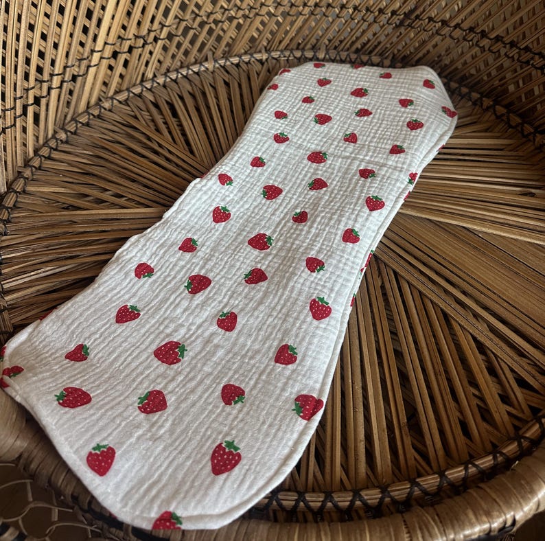 May include: A white baby bib with a pattern of red strawberries and green leaves. The bib is made of a soft, textured fabric and has a curved shape.