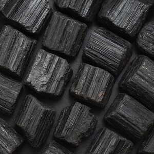 Puede incluir: Primer plano de cristales de turmalina negra en bruto. Los cristales son de un negro profundo y s&oacute;lido, con estr&iacute;as visibles y una textura natural rugosa. La disposici&oacute;n de los cristales llena el encuadre, creando una composici&oacute;n densa y rica en minerales.